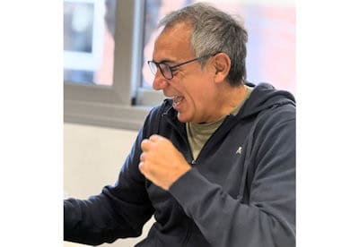 Man with gray hair and glasses laughing, wearing a dark zip-up jacket, gesturing with a clenched fist indoors.